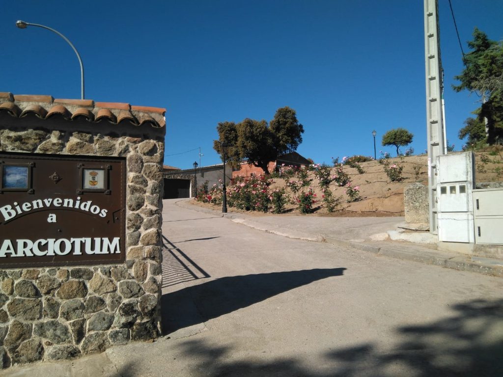 Cartel de piedra con la inscripción «Bienvenidos a Garciotum» a la entrada del pueblo, junto a la calle y un jardín con rosales