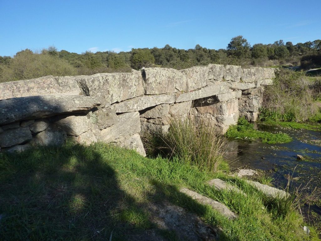 Puente de los Pilones: puente de grandes losas de granito sobre un arroyo, con hierba verde en la orilla y encinas al fondo