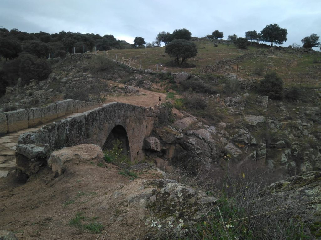 Puente Romano de Garciotum: puente de piedra de un solo ojo sobre un barranco rocoso, con un camino de tierra y encinas en la ladera