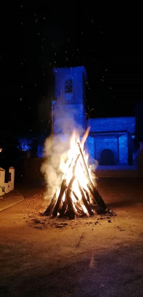 Gran hoguera encendida de noche en una calle, con la torre de la iglesia iluminada de azul al fondo